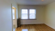 This single-floor space offers a warm and inviting atmosphere highlighted by timeless architectural character. The main living area features gleaming hardwood floors that reflect abundant natural light pouring in through a pair of large double-hung windows fitted with horizontal blinds. The windows provide views of the surrounding neighborhood, giving the space a connected, airy feel. A classic cast-iron radiator sits beneath the windows, a nod to traditional heating systems that add vintage charm and functional warmth to the room. Crown molding runs along the ceiling perimeter, adding a refined architectural detail that elevates the overall aesthetic. The walls are finished in a soft, neutral tone that complements the warm wood floors and bright trim work throughout. A doorway visible from the main room leads to an adjacent space, suggesting a practical and connected floor plan. The crisp white door frames and baseboards provide clean contrast against the light walls, reinforcing the space's classic character. Overall, the space combines period details with a bright, open ambiance, making it a well-lit and characterful environment suitable for a variety of uses.