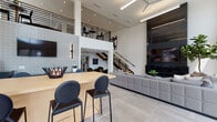 This contemporary two-story amenity space showcases striking architectural design defined by soaring double-height ceilings, floor-to-ceiling windows, and large-format porcelain tile flooring throughout. The main level opens into a grand, light-filled lounge area anchored by an expansive sectional sofa and a sleek wall-mounted fireplace clad in dark tile, creating a bold focal point. A communal high-top table with black leather bar chairs sits beneath towering windows that flood the space with natural light and offer views of an outdoor pool area. An adjacent lounge nook features a curved dark sectional sofa and wall-mounted television, providing a more intimate setting. The open-plan layout also incorporates a coffee and beverage station with warm wood cabinetry. A sculptural staircase with black metal railings and wood treads, accented by bold abstract artwork, connects to the second floor, where an open mezzanine overlooks the main living areas below. Upstairs, the game room is a standout feature, offering a full-size billiard table, generous seating, and panoramic windows with urban views. Throughout both levels, the design maintains a cohesive modern aesthetic with curated art pieces, lush indoor plants, and a refined neutral palette punctuated by bold accent furnishings.