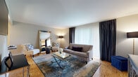 Located at 1 John F Kennedy Blvd, this thoughtfully designed single-floor residence combines classic architectural details with contemporary finishes throughout. The living room is a standout space, featuring warm parquet hardwood flooring, large windows dressed with elegant charcoal drapes that allow generous natural light, and a refined layout anchored by a glass-top coffee table and a full-length decorative mirror. Adjacent to the living area, a dedicated dining space accommodates an intimate glass-top round table surrounded by tufted upholstered chairs, accented by natural light and tasteful wall art. The kitchen is well-equipped with dark espresso cabinetry, granite countertops, stainless steel appliances including a gas range, built-in microwave, refrigerator, and dishwasher, all complemented by a classic subway tile backsplash and light tile flooring. The bedroom offers a serene retreat with plush carpeting, a tufted upholstered headboard, coordinating furniture, and a window framed by floor-length drapes. The full bathroom features floor-to-ceiling tiled walls in neutral tones, a soaking tub with shower, rich wood vanity cabinetry, and granite countertops. A dedicated laundry area with a full-size washer and dryer and overhead cabinetry adds everyday convenience.