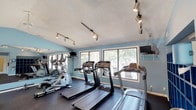 This fitness center is a thoughtfully designed, single-floor facility that offers a comprehensive workout environment. The space is painted in a calming light blue throughout, complemented by dark rubber flooring that provides durability and traction underfoot. Vaulted ceilings in the main exercise area add a sense of openness, while track lighting and ceiling fans keep the space well-lit and ventilated.

The main workout area is generously sized and lined with large windows on multiple walls, flooding the room with natural light and offering pleasant views of surrounding greenery and landscaping. A full-length wall mirror amplifies the sense of space and assists with form during exercise. Cardio equipment includes multiple treadmills and elliptical machines, while a dedicated strength area features a cable machine and a well-stocked dumbbell rack with a tiered weight selection.

A built-in blue cubby storage unit provides convenient space for personal belongings, and a water fountain is accessible nearby. Mounted televisions are positioned throughout for entertainment during workouts. A separate, quieter alcove houses the free weight station, offering a more focused training environment with a pass-through window connecting to an adjacent room. The facility also includes a clearly marked exit with direct outdoor access to the landscaped surroundings.