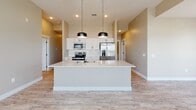 This well-appointed single-floor residence offers a thoughtfully designed open-concept layout that seamlessly connects the kitchen and living areas. The kitchen serves as the centerpiece of the home, showcasing a large white island with a matte black faucet and sink, crisp white shaker cabinetry, a classic subway tile backsplash, and a full suite of stainless steel appliances including a refrigerator with ice maker, electric range, and over-the-range microwave. Striking black pendant lights hang above the island, complementing the recessed lighting overhead. The soaring ceilings in the main living area amplify the sense of openness and airiness throughout. Light wood-look flooring extends consistently across all rooms, tying the space together with a warm, cohesive aesthetic. The hallway connects bedrooms and bathrooms efficiently, featuring recessed lighting and a programmable thermostat. The primary bathroom includes a white vanity with quartz countertop, an undermount sink with a matte black faucet, and a large mirror. A second bathroom offers a full tub-shower combination with built-in shelving and oil-rubbed bronze fixtures. A dedicated laundry room includes utility hookups, overhead shelving, and ample space for full-size appliances, adding significant practical value to this move-in ready home.