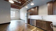 Located at 1600 O St in the heart of Lincoln, Nebraska, this one-bedroom, one-bathroom unit blends industrial character with modern finishes across a single open floor plan. The space makes a striking first impression with soaring ceilings accented by exposed wooden beams and open rafters, creating a loft-like atmosphere that feels remarkably spacious. Polished and stained concrete floors run throughout, reinforcing the industrial aesthetic while providing a durable, low-maintenance surface.

The kitchen is a standout feature, offering rich dark-stained cabinetry, granite countertops, a white subway tile backsplash, and a full suite of stainless steel appliances including a refrigerator, range, microwave, and dishwasher. Pendant lighting hangs above the kitchen area, complementing the exposed ceiling elements overhead.

The bedroom is accessed through a distinctive dark, solid-wood door that adds a bold design statement. Natural light enters through large windows, enhancing the open feel of the living areas. The bathroom features a clean, minimalist walk-in shower with a linear drain, white surround walls with a built-in niche, and wood-look tile flooring. Throughout the unit, dark baseboards and door frames provide sharp contrast against the light-gray walls, tying the overall industrial-modern aesthetic together cohesively.