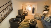 This two-floor residence offers a welcoming and functional layout designed for everyday comfort. The main living area features neutral-toned walls and plush carpeted flooring, creating a warm and inviting atmosphere. A ceiling fan with integrated lighting adds both practical ventilation and a classic finishing touch to the room. The open floor plan allows the living space to flow seamlessly into a dedicated dining area, which comfortably accommodates a bistro-style table and chairs — ideal for casual meals or intimate gatherings. Just beyond, the kitchen is visible and open to the main living space, featuring standard appliances including a full-size refrigerator, and benefits from natural light streaming in through a window above the counter. The overall color palette throughout the main level is soft and neutral, with warm beige tones that lend a cohesive, lived-in feel. Staircase access to the second floor is visible from the living area, offering a clear sense of the home's two-story nature. The layout balances openness with defined zones for living, dining, and cooking, making it a practical and comfortable space suitable for a range of lifestyles.