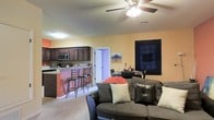This single-floor space offers an open-concept layout that seamlessly connects the living, dining, and kitchen areas into one cohesive and functional environment. The living room is anchored by a plush dark leather sofa accented with decorative pillows, creating a comfortable and welcoming atmosphere. A ceiling fan with an integrated light fixture overhead adds both practicality and a finishing touch to the space.

The kitchen is well-appointed with dark wood cabinetry, a tile backsplash, and includes modern appliances such as a microwave and oven. A raised breakfast bar with bar-height seating provides a casual dining option, perfect for everyday meals or entertaining guests. Adjacent to the kitchen, a dedicated dining area offers additional seating for more formal gatherings.

The warm peach and terracotta wall tones lend a cozy, inviting character throughout, complemented by light-colored flooring that brightens the overall space. A doorway visible from the main living area suggests additional rooms beyond, adding depth and functionality to the floor plan. The layout is efficient and thoughtfully designed, making excellent use of the available space while maintaining an open and airy feel throughout.
