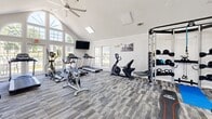 Located at 99 Ascension Dr in Asheville, NC, this well-appointed fitness center offers a spacious, open layout designed to support a wide range of workout routines. The space is immediately distinguished by its soaring vaulted ceilings, which lend an airy, expansive feel, and are accented by skylights that pour natural light throughout the room. Large arched and rectangular windows line the walls, framing views of the surrounding greenery and further brightening the interior.

The flooring consists of durable, patterned carpet tiles in a neutral gray tone, providing both comfort and traction underfoot. Cardio equipment is well-represented, with multiple treadmills, stationary bikes, a spin bike, and an elliptical trainer arranged for easy access. Strength training is equally supported, with a multi-station cable machine, a dedicated weight bench, a tiered dumbbell rack, and kettlebells. Stability balls and exercise mats offer options for flexibility and functional training.

A wall-mounted flat-screen television is centrally positioned for viewing during workouts, while a ceiling fan contributes to air circulation. A water cooler station and adjacent rooms — visible through open doorways — suggest additional amenity spaces nearby. Motivational wall art and crisp white walls with crown molding complete the clean, welcoming aesthetic of this community fitness facility.