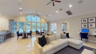 This single-story space is designed with an open-concept layout that seamlessly connects multiple functional areas beneath soaring vaulted ceilings. Large arched windows and French doors flood the interior with natural light throughout, while offering views of a outdoor pool and surrounding mature trees.

The main gathering area is anchored by a tile-surround fireplace with a wall-mounted television above, flanked by arched windows and accented by ceiling fans. Comfortable seating arrangements and a neutral color palette with navy blue accents create an inviting, polished atmosphere. An adjoining lounge area features a built-in workstation with computer terminals and gallery-style framed artwork, making it well-suited for both relaxation and productivity.

The dining area benefits from expansive floor-to-ceiling arched windows and direct access to the outdoor patio via French doors, providing an airy, sun-drenched setting. The kitchen is equipped with rich dark-wood cabinetry, granite countertops, a stainless steel refrigerator, and a breakfast bar with seating.

The entryway features a decorative console table, textured walls, and a pass-through window into a secondary office area. Throughout, polished tile flooring, recessed lighting, and crown molding contribute to a refined, cohesive aesthetic.