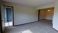 This single-floor space presents a clean and functional layout designed with simplicity in mind. The main living area is spacious and open, featuring wall-to-wall carpet in a neutral gray tone that adds a soft, understated feel underfoot. The walls are painted in a light, off-white hue, contributing to a bright and airy atmosphere throughout the interior.

The room features standard-height ceilings and smooth, plain wall surfaces, giving the space a blank-canvas quality that is well-suited for personalization. Dark baseboard trim runs along the perimeter of the room, providing a subtle contrast to the light walls and flooring. Electrical outlets are evenly distributed along the walls, offering practical placement for furniture and appliances.

A partial wall division is visible toward the back of the main area, leading into a connected secondary space. This adjoining room appears to offer additional functional square footage, with a baseboard heating unit positioned along the far wall — a practical feature for climate control. The overall layout flows naturally between areas, making the space feel cohesive and easy to navigate. The unit offers a straightforward, move-in-ready environment with essential features in place.