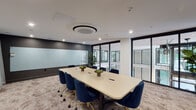 Located at 375 Camino De La Reina in San Diego, CA 92108, this sophisticated two-floor office space delivers a polished, modern work environment designed for productivity and collaboration. The interior is defined by a refined design aesthetic, combining dark charcoal accent walls with crisp white surfaces and warm, textured carpeting that adds depth and comfort underfoot.

The conference room serves as a centerpiece of the space, featuring a spacious rectangular table surrounded by navy blue velvet chairs with gold-toned casters — a striking combination that balances professionalism with contemporary style. A large frosted glass whiteboard spans one wall, providing an expansive surface for presentations and brainstorming. A wall-mounted display screen adds integrated media capability, while a circular ceiling-mounted HVAC unit and recessed lighting ensure a comfortable and well-lit atmosphere.

Floor-to-ceiling black-framed glass partition walls allow for visual connectivity between spaces, flooding the interior with borrowed light and offering views across both floors. Lush indoor plants, including a prominent fiddle-leaf fig, introduce organic warmth throughout. The open sightlines between levels reinforce the building's airy, collaborative character, making this a highly functional and visually impressive professional environment.