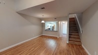This well-appointed 1-bedroom, 1.5-bathroom two-story residence offers a generous and thoughtfully designed layout across two floors. The main level features an expansive open-concept living and dining area finished with warm-toned hardwood flooring that extends throughout, creating a bright and cohesive atmosphere. Neutral, soft gray walls complement the light wood tones, lending the space a clean and modern aesthetic.

A pass-through kitchen window connects the living area to the kitchen, which is outfitted with white cabinetry, dark countertops, and stainless steel appliances including a full-size refrigerator. A pendant light fixture anchors the dining area, adding a subtle decorative touch. A staircase with wood-toned treads leads to the upper level, where the bedroom and additional bathroom are located, providing a comfortable separation between living and sleeping spaces.

The half bathroom on the main level adds everyday convenience, making the layout practical for both daily living and entertaining. The entry area is accessible from the main floor, completing a functional and well-organized floor plan. Overall, this two-story residence balances spaciousness with a warm, inviting ambiance suited for comfortable everyday living.
