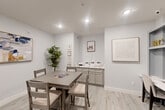 Located at 1301 Rudel Drive in Tomball, Texas, this single-room space offers a well-appointed and versatile layout that seamlessly blends dining and workspace functionality. The room is finished with light wood-look flooring throughout, crisp white walls, and recessed lighting that creates a bright, clean ambiance.

The dining area anchors the center of the room, featuring a square table with four cushioned chairs — an ideal setting for small meetings or casual gatherings. Along one wall, sage-toned cabinetry with a light countertop provides ample storage and workspace for equipment or supplies, making it a practical utility zone.

A standout feature is the built-in workstation along the adjacent wall, where a marble-style surface countertop spans the width of the space, flanked by open shelving above. This area comfortably accommodates two seated workspaces, with cubbies providing display and storage space. Framed artwork adorns the walls, adding a polished, professional aesthetic throughout.

A private entry door with slatted blinds allows for natural light control, while the overall neutral color palette lends the space a calm and focused atmosphere. This thoughtfully designed room is well-suited for use as a shared office, creative studio, or meeting space.