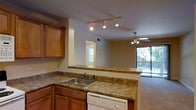 Located at 820 McMillan Avenue in Wilmington, North Carolina, this single-floor residence offers a well-designed open-concept layout that seamlessly connects the kitchen and living areas. The kitchen is equipped with warm honey-toned cabinetry, stone-patterned countertops, a stainless steel sink, and a built-in dishwasher. A peninsula counter serves as a natural divider between the kitchen and the adjoining living space, making it ideal for casual dining or entertaining. Track lighting above the kitchen provides focused illumination, while a ceiling fan in the living area ensures year-round comfort. Natural light fills the space through multiple windows, including a large sliding glass door that leads to a private balcony with views of lush outdoor greenery. The carpeted living area creates a comfortable and inviting atmosphere. A hallway visible from the main living space suggests additional rooms beyond, contributing to the overall functional flow of the floor plan. The combination of practical finishes and an open, airy layout makes this a comfortable and versatile living space.