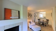 This single-floor residence offers a thoughtfully arranged layout that seamlessly connects the living, dining, and kitchen areas into a cohesive, comfortable living environment. The living room serves as the heart of the space, anchored by a classic white-tiled fireplace with a dark hearth that adds a warm, inviting focal point. Neutral walls and light carpeting throughout lend the interior a clean, airy feel, while framed artwork adds subtle character to the walls.

A half-wall architectural detail divides the living room from the dining area, providing a sense of separation while maintaining an open flow between the two spaces. The dining area accommodates a round table with traditional-style chairs and is illuminated by a pendant light fixture, creating an intimate setting for meals. Natural light filters in through a window near the dining space, brightening the area and offering a glimpse into the adjacent kitchen.

The kitchen is visible in the background, featuring light cabinetry and counter space, and is conveniently accessible from the dining zone. A hallway beyond the living area suggests additional rooms, reinforcing the functional and well-organized nature of the floor plan. Overall, the space presents a harmonious blend of classic design elements and practical layout, suited for comfortable everyday living.
