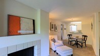 This single-floor residence offers a thoughtfully arranged layout that seamlessly connects the living, dining, and kitchen areas into a cohesive, comfortable living environment. The living room serves as the heart of the space, anchored by a classic white-tiled fireplace with a dark hearth that adds a warm, inviting focal point. Neutral walls and light carpeting throughout lend the interior a clean, airy feel, while framed artwork adds subtle character to the walls.

A half-wall architectural detail divides the living room from the dining area, providing a sense of separation while maintaining an open flow between the two spaces. The dining area accommodates a round table with traditional-style chairs and is illuminated by a pendant light fixture, creating an intimate setting for meals. Natural light filters in through a window near the dining space, brightening the area and offering a glimpse into the adjacent kitchen.

The kitchen is visible in the background, featuring light cabinetry and counter space, and is conveniently accessible from the dining zone. A hallway beyond the living area suggests additional rooms, reinforcing the functional and well-organized nature of the floor plan. Overall, the space presents a harmonious blend of classic design elements and practical layout, suited for comfortable everyday living.