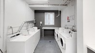 This shared laundry facility is designed with practicality and efficiency in mind. The room is finished with crisp white walls and smooth concrete floors, creating a clean, utilitarian aesthetic throughout. Exposed white pipes and silver ventilation ducts run along the ceiling, adding an industrial character to the space.

Multiple top-load washing machines are arranged along one wall, while a row of front-load washers and dryers lines the opposite side, providing ample capacity for simultaneous use. A central folding counter with built-in storage below offers a dedicated workspace for handling laundry. An additional built-in countertop with overhead cabinet storage is available near the entrance area, providing extra utility space.

A small window on the painted grey brick accent wall allows in a modest amount of natural light. Fluorescent ceiling fixtures ensure the room remains well-lit throughout. Seating is available along the walls, offering a comfortable place to wait. Informational signage is posted throughout the room for user guidance.

Access to the facility is provided through a wood-framed door with a clearly marked exit sign, connecting to a stairwell. A security camera is also present, contributing to the overall safety of the space.