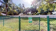 This designated pet area is a dedicated outdoor space exclusively for residents and their pets. Enclosed by a green chain-link fence with a gated entry, the area provides a safe and contained environment for dogs and cats to roam freely. A clearly posted sign at the entrance reminds residents to clean up after their pets, reinforcing a clean and well-maintained environment. The ground is covered in grass, offering a natural surface for pets to enjoy. The space is framed by a white perimeter wall and surrounded by rich tropical landscaping, including towering palm trees and dense mature foliage that create a lush, shaded backdrop. The abundance of greenery lends the area a tranquil, park-like atmosphere. Bright natural light filters through the canopy, enhancing the open and airy feel of the space. This pet area reflects a thoughtful amenity designed to accommodate the needs of pet-owning residents while maintaining an organized and pleasant outdoor environment.