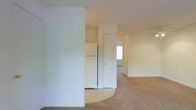 This single-floor unit features a straightforward, functional layout with a clear division between the living and kitchen areas. The living space is finished with wall-to-wall carpet in a neutral beige tone, providing a warm and understated aesthetic that carries throughout the main areas. A partial wall with a curved base acts as a subtle architectural divider, separating the carpeted living zone from the kitchen area, which transitions to light-toned tile flooring.

The kitchen is equipped with a full-size white refrigerator and includes a closet or pantry with a white-paneled door, offering practical storage. The walls throughout are painted in a clean, off-white palette, contributing to a bright and airy feel. Natural light filters in through a window visible toward the rear of the unit, illuminating the adjoining space and adding depth to the overall layout.

The design is simple and utilitarian, with standard ceiling heights and minimal architectural ornamentation. The neutral finishes and open sightlines between rooms make the space feel cohesive and easy to furnish, suited for everyday residential use.