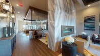 This single-story clubhouse is a well-appointed amenity space designed with upscale finishes and diverse social areas throughout. The entry lounge serves as a striking focal point, featuring a dramatic floor-to-ceiling marble-clad column housing a sleek linear fireplace, complemented by richly veined stone wall panels, soaring ceilings, and curated furnishings including leather sofas and sculptural accent tables beneath an oversized pendant light.

Adjacent seating areas transition seamlessly across wood-look tile flooring, with patterned area rugs defining distinct zones. A dedicated dining area accommodates a large communal table surrounded by mixed seating and illuminated by statement globe chandeliers in brass, with expansive glass doors drawing in natural light and outdoor views.

The galley-style kitchen features dual runs of dark cabinetry topped with quartz countertops, built-in appliances, a mirrored tile backsplash, and pendant lighting, providing ample space for catering or group gatherings.

A games and entertainment area includes a shuffleboard table, casual dining seating, mounted television, and a glass-enclosed golf simulator bay alongside a billiards table. A separate lounge with tall windows and plush leather sectionals offers a quieter retreat with abundant natural light.