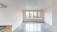 This contemporary single-floor unit showcases a clean, modern aesthetic defined by polished concrete floors that run seamlessly throughout the entire space, reflecting natural light and lending an industrial-chic character to every room. The open-concept layout connects the living area and kitchen, creating a cohesive and functional living environment.

The kitchen is efficiently designed with warm wood cabinetry, a dark metal backsplash, concrete countertops, stainless steel appliances including a refrigerator and dishwasher, and a stainless steel range hood as a standout focal point. Open shelving above the cabinetry adds both storage and visual interest. A wall-mounted split air conditioning unit serves the main living area, ensuring year-round comfort.

The generously proportioned living area is bathed in natural light from a wide, multi-pane window with warm wood framing, offering elevated views of the surrounding urban cityscape and neighboring buildings framed by tree canopies. The crisp white walls enhance the brightness of the space.

The bathroom features classic white subway tile surrounding a full bathtub and shower, complemented by a wood vanity with a concrete countertop and integrated sink, maintaining the cohesive material palette found throughout the unit. A well-lit entryway corridor with tall cabinetry provides ample storage upon entry.
