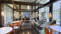 This sophisticated lounge space is defined by its dramatic double-height ceilings, accented by prominent dark structural beams that create a striking geometric framework overhead. The open-plan layout is flooded with natural light streaming through expansive floor-to-ceiling windows along the perimeter, offering views of lush greenery outside. The rich, dark hardwood flooring anchors the space and contrasts elegantly with the lighter wall tones and furnishings throughout. Multiple distinct seating areas are arranged throughout the room, including a central lounge zone centered around a wall-mounted flat-screen television, surrounded by contemporary sofas, accent chairs, and floor lamps. A casual dining area near the front features a round white table paired with translucent amber chairs, adding a playful pop of color. Additional seating clusters with modern swivel chairs provide versatile gathering spots. Pendant lighting in warm tones descends from the ceiling framework, complementing the ambient glow of strategically placed floor lamps. Contemporary artwork adorns the walls, enhancing the polished, upscale atmosphere. The overall ambiance is open, modern, and welcoming, designed for both social interaction and relaxed lounging.