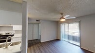 This single-floor unit offers a well-organized layout with consistent dark hardwood-style flooring flowing throughout the living spaces. The living room is a comfortable, open area featuring a ceiling fan with integrated lighting and a sliding glass door flanked by vertical blinds, providing access to an outdoor patio and allowing natural light to fill the room. A closet with bifold doors adds convenient built-in storage to the space.

The kitchen is designed for functionality and visual appeal, featuring white cabinetry, stainless steel appliances including a refrigerator and electric range, a double-basin sink, and a light-colored countertop that extends to form a breakfast bar or pass-through, connecting the kitchen to the adjacent dining area. A pendant light fixture illuminates the dining space, which benefits from the open sightlines to the kitchen.

A dedicated bedroom offers a quiet retreat, complete with textured ceilings consistent throughout the unit and ample wall space. The bathroom is clean and straightforward, equipped with a full tub-and-shower combination featuring white square tile surround, brushed nickel fixtures and accessories, and coordinating hardwood-style flooring. Overall, the unit presents a neutral, modern palette with grey walls, white trim, and warm flooring tones.