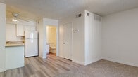 This single-floor unit offers a straightforward, functional layout designed for comfortable everyday living. The main living area is generously proportioned, featuring wall-to-wall carpet in a neutral beige tone that adds warmth underfoot. A window fitted with vertical blinds allows natural light to filter into the space while offering privacy, and a front entry door provides direct access to the unit. The textured ceiling runs throughout, lending a consistent finish across all areas.

The kitchen is efficiently arranged along one wall, featuring white cabinetry with ample storage both above and below the counter. A double-basin stainless steel sink is set into a tan laminate countertop, and a white refrigerator is positioned nearby. The kitchen floor transitions to a light-toned hardwood-style surface, clearly delineating it from the carpeted living space.

Adjacent to the kitchen area is a closet and access to the bathroom, which includes a toilet and vanity with white cabinetry, continuing the clean, consistent aesthetic found throughout the unit. HVAC vents are visible on the walls, ensuring climate control throughout the space. Overall, the unit maintains a clean, neutral palette of whites and beiges, creating a bright and move-in ready environment.
