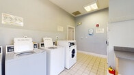 Located at 150 Carrington Park Drive in Gainesville, Georgia, this community laundry facility is a well-maintained, single-floor space designed for convenient and comfortable use. The room is finished with clean light-colored tile flooring and soft blue-gray walls, creating a calm and functional atmosphere throughout.

The laundry area is equipped with multiple top-loading washers and at least one front-loading washer and dryer unit, all fitted with card-operated payment systems. Instructional signage is posted along the walls to assist users with machine operation. A built-in folding counter with an overhead wire shelf provides a dedicated space for sorting and folding laundry, complemented by a cushioned bench nearby for added convenience.

Natural light fills the space generously through large multi-pane windows and a set of glass-panel French doors that open to an outdoor courtyard area, offering views of surrounding greenery and a pool area. Overhead fluorescent lighting ensures the space remains well-lit at all times. A hallway connects to an adjacent room with light wood-style flooring, accessible through a glass-pane interior door. Safety features include a fire extinguisher and a fire alarm mounted on the wall. The overall design is practical, clean, and welcoming for everyday community use.