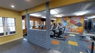 This fitness center is designed to provide an motivating and functional workout environment. The space is defined by bold mustard-yellow accent walls that inject energy into the room, complemented by a large, colorful mural depicting an athletic figure in motion — a striking focal point that reinforces the space's active purpose.

The flooring consists of patterned commercial carpet tiles in grey, orange, and yellow, adding visual interest while providing a practical, durable surface underfoot. Structural columns with decorative textured paneling featuring an organic, pebble-like pattern serve as both architectural elements and subtle room dividers, creating distinct zones within the open layout.

Natural light filters in through several windows along one wall, brightening the space alongside overhead recessed and flush-mount lighting fixtures that ensure consistent illumination throughout. The cardio equipment section is well-stocked with elliptical trainers and treadmills, arranged to allow comfortable spacing between machines. Mirrors along the walls visually expand the space and allow users to monitor their form during workouts. Overall, the fitness center balances practicality with an aesthetically engaging design.