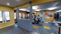 This fitness center is designed to provide an motivating and functional workout environment. The space is defined by bold mustard-yellow accent walls that inject energy into the room, complemented by a large, colorful mural depicting an athletic figure in motion — a striking focal point that reinforces the space's active purpose.

The flooring consists of patterned commercial carpet tiles in grey, orange, and yellow, adding visual interest while providing a practical, durable surface underfoot. Structural columns with decorative textured paneling featuring an organic, pebble-like pattern serve as both architectural elements and subtle room dividers, creating distinct zones within the open layout.

Natural light filters in through several windows along one wall, brightening the space alongside overhead recessed and flush-mount lighting fixtures that ensure consistent illumination throughout. The cardio equipment section is well-stocked with elliptical trainers and treadmills, arranged to allow comfortable spacing between machines. Mirrors along the walls visually expand the space and allow users to monitor their form during workouts. Overall, the fitness center balances practicality with an aesthetically engaging design.