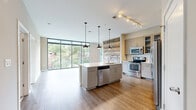 This well-appointed 3-bedroom, 2-bathroom residence offers a thoughtfully designed single-floor layout with a clean, contemporary aesthetic. The open-concept kitchen and living area serves as the heart of the home, anchored by a central island with a built-in sink and dishwasher, complemented by stainless steel appliances, light wood cabinetry with glass-front upper cabinets, white subway tile backsplash, and warm hardwood-style flooring. A striking reclaimed wood accent wall adds texture and character to the space. Industrial-style pendant lights hang above the island, while a track lighting system illuminates the kitchen zone. Floor-to-ceiling windows flood the living area with abundant natural light and frame lush treetop views of the surrounding neighborhood. The spacious primary bedroom includes direct access to a private balcony through sliding glass doors, offering pleasant outdoor views. Additional bedrooms feature large windows with neighborhood views and durable wood-style flooring. The bathrooms are finished with large-format tile flooring, modern vanities, and sleek hardware. A dedicated walk-in closet with wire shelving systems provides generous storage. Throughout the residence, neutral warm tones and consistent flooring create a cohesive, modern ambiance.
