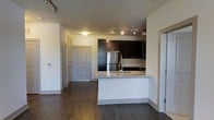 This single-floor unit showcases a thoughtfully designed open-concept layout that seamlessly connects the kitchen and living areas. The kitchen serves as a central feature, anchored by a curved peninsula with a granite countertop that doubles as a breakfast bar. Dark espresso cabinetry contrasts elegantly against the lighter walls and ceiling, while stainless steel appliances, including a full-sized refrigerator, add a polished, modern touch. A track lighting system mounted to the ceiling provides focused illumination across the kitchen workspace, complementing the ambient overhead lighting in the adjoining area. Rich, dark hardwood flooring runs throughout the space, adding warmth and continuity to the open layout. The entryway area is well-defined, featuring a paneled front door alongside additional doors that suggest closet storage, offering practical organization. The neutral wall palette keeps the space feeling bright and versatile. Overall, the unit presents a clean, contemporary aesthetic with functional design elements that balance style and everyday practicality.