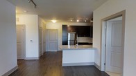 This single-floor unit showcases a thoughtfully designed open-concept layout that seamlessly connects the kitchen and living areas. The kitchen serves as a central feature, anchored by a curved peninsula with a granite countertop that doubles as a breakfast bar. Dark espresso cabinetry contrasts elegantly against the lighter walls and ceiling, while stainless steel appliances, including a full-sized refrigerator, add a polished, modern touch. A track lighting system mounted to the ceiling provides focused illumination across the kitchen workspace, complementing the ambient overhead lighting in the adjoining area. Rich, dark hardwood flooring runs throughout the space, adding warmth and continuity to the open layout. The entryway area is well-defined, featuring a paneled front door alongside additional doors that suggest closet storage, offering practical organization. The neutral wall palette keeps the space feeling bright and versatile. Overall, the unit presents a clean, contemporary aesthetic with functional design elements that balance style and everyday practicality.