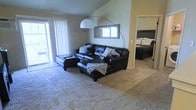 This single-story residence features an open-concept living area distinguished by vaulted ceilings that lend a sense of spaciousness to the interior. The living room is anchored by plush, neutral-toned carpeting that extends throughout the space, creating a warm and cohesive aesthetic. A large sliding glass door fitted with vertical blinds fills the room with natural light while providing direct access to an outdoor area, offering a bright and airy atmosphere.

The living area accommodates generous seating, as evidenced by the substantial sectional sofa arrangement, and is complemented by a striking arc floor lamp that serves as a functional design focal point. A framed mirror mounted on the wall above the sofa enhances the sense of depth in the room.

An open doorway connects the living area to an adjacent bedroom, which features dark-toned furniture and maintains the same carpeted flooring, reinforcing the continuity of the interior design. The layout promotes an easy flow between living and sleeping spaces, making the residence feel well-connected and efficiently arranged throughout its single level.