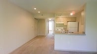 This single-floor space features an open-concept design that integrates the living area and kitchen into one cohesive layout. The living area is finished with neutral-toned carpeting that extends throughout, contributing to a warm and understated atmosphere. Recessed lighting and track lighting are installed overhead, providing consistent illumination across both zones.

The kitchen is positioned along one side of the open floor plan and includes light wood cabinetry offering ample storage space. A white electric range, a refrigerator, and an under-cabinet range hood are among the appliances present. A peninsula with a dual-basin sink and countertop seating separates the kitchen from the living area, serving as both a functional workspace and a casual dining option.

Multiple doors are visible along the rear wall of the living area, suggesting access to additional rooms such as bedrooms or storage closets. A hallway leads toward a brightened area, likely a bedroom or bathroom with natural light. HVAC vents are visible in the ceiling, indicating a central air system. Overall, the space presents a clean, practical layout suited for comfortable day-to-day living.