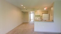 This single-floor space features an open-concept design that integrates the living area and kitchen into one cohesive layout. The living area is finished with neutral-toned carpeting that extends throughout, contributing to a warm and understated atmosphere. Recessed lighting and track lighting are installed overhead, providing consistent illumination across both zones.

The kitchen is positioned along one side of the open floor plan and includes light wood cabinetry offering ample storage space. A white electric range, a refrigerator, and an under-cabinet range hood are among the appliances present. A peninsula with a dual-basin sink and countertop seating separates the kitchen from the living area, serving as both a functional workspace and a casual dining option.

Multiple doors are visible along the rear wall of the living area, suggesting access to additional rooms such as bedrooms or storage closets. A hallway leads toward a brightened area, likely a bedroom or bathroom with natural light. HVAC vents are visible in the ceiling, indicating a central air system. Overall, the space presents a clean, practical layout suited for comfortable day-to-day living.