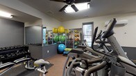 This single-level fitness center is thoughtfully designed to support a wide range of workout routines. The space is finished with durable cork flooring throughout, complemented by protective rubber mats positioned beneath the equipment. Two-tone walls — light gray above and darker gray wainscoting below — give the room a clean, structured aesthetic, while large wall mirrors along one side visually expand the space and aid in form correction during exercise.

Cardio equipment is a focal point of the room, with Matrix-brand treadmills and elliptical trainers positioned near the windows, allowing natural light to filter in during workouts. A wall-mounted flat-screen television provides entertainment, while a wall-mounted mini-split HVAC unit ensures climate comfort year-round. A ceiling fan adds additional air circulation overhead.

The functional training area features a well-organized rack of stability balls in multiple sizes, resistance accessories, and foam rollers, offering ample options for stretching and core work. A multi-function cable machine and a stationary bike round out the equipment selection. A secured entry door with a window panel connects the fitness center to an adjacent common area, and multiple windows provide views to the surrounding exterior, keeping the space bright and open.