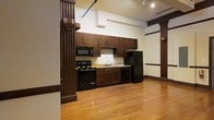This single-floor space combines classic architectural details with a functional, open layout. The kitchen is a standout feature, showcasing rich, dark wood cabinetry that extends to the upper walls, providing ample storage. Black appliances — including a gas range, built-in microwave, and refrigerator — complement the cabinetry for a cohesive, bold aesthetic. A sink with a clean, simple design is centrally positioned along the countertop, contributing to the kitchen's practical flow.

Warm, honey-toned hardwood floors span the open living area, lending a sense of continuity and spaciousness throughout the space. A defining architectural element is the prominent fluted wood columns, which frame the room with a sense of character and craftsmanship reminiscent of historic building design. Additional decorative wood molding and trim details reinforce this classic aesthetic.

Overhead, a track lighting system mounted to the ceiling provides flexible, directed illumination across the space. The elevated ceiling height enhances the sense of openness and volume, giving the interior an airy quality. The combination of rich woodwork, open sightlines, and industrial-style lighting creates a space that balances historic charm with modern livability.