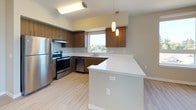 This single-floor residence offers a thoughtfully designed layout that balances functionality and modern aesthetics. The kitchen serves as a central highlight, featuring rich warm-toned wood cabinetry, crisp white countertops, and a full suite of stainless steel appliances including a refrigerator, range, and dishwasher. A large window above the sink frames picturesque views of surrounding trees and hillside, filling the space with natural light. An extended white peninsula provides additional prep space and serves as a natural divider between the kitchen and the adjoining living area. Light-toned hardwood-style flooring runs throughout the main living and kitchen areas, contributing to a clean, contemporary feel. The bedroom is generously proportioned, finished with soft neutral carpet, and features a wide window that captures the same scenic outdoor views, offering a bright and tranquil retreat. A dedicated laundry area houses a full-size front-loading washer and dryer, adding a practical and convenient amenity within the unit. The bathroom is clean and functional, complete with a full-size soaking tub and shower combination finished in crisp white surround. Throughout the space, neutral wall tones and consistent modern finishes create a cohesive and inviting atmosphere well-suited for comfortable everyday living.