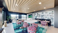 This rooftop amenity level combines a stylishly designed indoor lounge with a generous open-air terrace, offering a well-rounded social environment. Inside, the lounge features a warm slatted wood ceiling with recessed lighting that contrasts beautifully against dark accent walls. A bold, large-scale botanical mural anchors one wall, while vibrant teal-toned patterned carpeting adds energy to the space. Seating is varied and eclectic, including round white pedestal tables with colorful upholstered bar stools and intimate banquette-style booth seating. A foosball table and mounted flat-screen television add recreational appeal, making the interior well-suited for both casual gatherings and organized events. Large windows along the perimeter allow abundant natural light and connect the interior visually to the surrounding cityscape. The outdoor terrace extends the living space considerably, featuring multiple distinct zones: a lounge area with cushioned chairs surrounding a rectangular fire pit, a dining area with metal tables and teal wire chairs, and a grilling station with built-in cabinetry. Overhead string lights create an inviting atmosphere after dark, while unobstructed panoramic views of the surrounding urban landscape make the terrace a standout feature of this elevated amenity space.