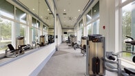 Located on NE 79th Street in Miami, FL, this well-appointed fitness center offers a comprehensive workout environment across a single, thoughtfully designed floor. The facility is characterized by soaring ceilings, large floor-to-ceiling windows that flood the space with natural light, and views of the surrounding lush greenery — creating an open, energizing atmosphere throughout.

The main cardio area is generously sized, housing a row of treadmills, elliptical machines, and stationary bikes positioned along the windowed walls to take advantage of the outdoor views. Ceiling fans overhead contribute to a comfortable workout environment. Wall-mounted televisions are positioned throughout for entertainment during exercise sessions.

A dedicated strength training corridor features a full lineup of Precor selectorized machines arranged along one wall, with large mirrors lining the opposite side to maximize the sense of space and allow for proper form checks. This section flows seamlessly into a functional training area stocked with free weights, dumbbells, barbells, benches, stability balls, and medicine balls — catering to a wide range of training styles.

The facility also includes a water fountain, mechanical and administrative support rooms, and clearly marked emergency exits. The neutral gray carpeting, white walls with blue accent trim, and recessed lighting combine to give the space a clean, professional aesthetic throughout.