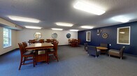 This single-floor space combines a spacious communal gathering room with a functional kitchen area, offering a well-rounded environment suitable for group activities and shared dining. The main gathering room features blue-gray carpet throughout and is illuminated by multiple recessed rectangular fluorescent light panels set into a textured ceiling, providing consistent, even lighting across the space. A bold navy blue accent wall serves as a focal point, adorned with nautical-themed decorations including a ship's wheel, an anchor, and circular porthole-style mirrors that echo a maritime aesthetic. The remaining walls are finished in neutral white and also feature round decorative mirrors, contributing to a cohesive design theme. Seating is arranged across several sturdy wooden dining tables with matching mission-style chairs, accommodating groups of various sizes. A separate lounge area includes upholstered gray armchairs arranged around a round wooden table, offering a more relaxed setting. Natural light enters through a window near the exit door. The adjacent kitchen area features warm honey-toned wood cabinetry, hardwood-style flooring, a white electric range, microwave, full-size refrigerator, and a sink, making the space fully equipped for preparing and serving meals.