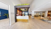 The Stonecrest Drive Clubhouse in Birmingham, Alabama offers a thoughtfully designed, single-floor community facility that blends modern aesthetics with practical amenities. The entrance greets visitors with a striking reception area anchored by a marble-topped desk with warm wood paneling, framed by a bold architectural archway adorned with lush trailing greenery. Large-format tile flooring extends throughout the main areas, lending a clean and contemporary feel.

Adjacent to the lobby, a co-working and lounge area provides both collaborative and focused work options. A long communal wooden table is surrounded by rattan-accented chairs, while dedicated workstations line a blue textured accent wall beneath soaring ceilings. Abundant natural light pours through generous windows, and curated artwork adds vibrant personality throughout.

A comfortable lounge area features a mounted flat-screen TV, stylish seating, and large windows framing views of the surrounding greenery. A glass-walled private conference room offers a quiet, professional meeting space accessible directly from the main lounge.

The kitchenette is outfitted with butcher block countertops, bold blue cabinetry, a stainless steel sink, open shelving, and a coffee station. The expansive fitness center is a standout feature, boasting vaulted ceilings, large windows, a full array of strength and cardio equipment, mirrored walls, and vibrant geometric flooring — all accented by a teal color scheme.