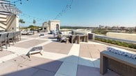 Located at 1100 S Main St in Greenville, South Carolina, this rooftop amenity deck offers an expansive outdoor experience with a diverse range of recreational and social features. The space is finished with large-format pavers throughout, providing a clean, modern aesthetic that unifies the various activity zones.

At the heart of the deck, comfortable wicker lounge seating arrangements are positioned to take full advantage of panoramic views of the Greenville skyline and the rolling Blue Ridge foothills beyond. Glass railings along the perimeter ensure unobstructed sightlines from every angle. String lights draped overhead add a welcoming ambiance for evening gatherings.

A dedicated outdoor bar area sits beneath a modern pergola, complete with bar-height seating and what appears to be a built-in grill or kitchen setup, making it ideal for entertaining. An outdoor television mounted on a white accent wall nearby enhances the al fresco viewing experience. A wood-burning fireplace provides a cozy focal point on one end of the deck.

For recreation, a well-maintained putting green is tucked into a dedicated corner of the rooftop, enclosed by frosted panel fencing and accented with young plantings. Cornhole boards are also available for casual outdoor play. Together, these amenities create a dynamic, resort-style rooftop destination in the heart of downtown Greenville.