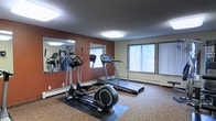 This single-floor fitness room is designed to provide a functional and motivating workout environment. The space is outfitted with a selection of cardio equipment, including a treadmill and an elliptical cross-trainer, both positioned on protective rubber floor mats over carpeted flooring. A large wall-mounted mirror spans a significant portion of one side of the room, visually expanding the space and allowing users to monitor their form during exercise. The accent wall, painted in a deep terracotta red, adds a bold, energizing contrast to the neutral tones of the surrounding walls and ceiling. Natural light filters into the room through two generously sized windows fitted with vertical blinds, offering a pleasant connection to the outdoors while maintaining privacy. Overhead, flush-mounted ceiling lights provide consistent, even illumination throughout the space. A baseboard heating unit runs along the exterior wall beneath the windows, ensuring year-round comfort. The textured ceiling and carpeted floors contribute to sound absorption, making the environment conducive to focused exercise. Overall, the fitness room is a practical and well-appointed amenity space.
