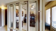 This single-floor professional office space offers a polished and well-appointed environment designed for productivity and a welcoming atmosphere. The office entry is defined by elegant glass-paneled French-style doors with dark brown trim, providing a clear sightline into the workspace while maintaining a sense of distinction and privacy. A gold placard labeled "Office" marks the entrance, adding a refined, professional touch.

Inside, the space features light tile flooring that complements the warm, neutral wall tones throughout. A ceiling fan with integrated lighting provides both air circulation and ambient illumination, supplemented by recessed lighting that brightens the interior evenly. Dark brown cabinetry and shelving offer substantial storage and display space, maintaining a cohesive, rich color palette throughout.

Decorative elements include tall decorative vases displayed atop the cabinetry, a bold floral wall art piece in warm amber and brown tones, and a patterned area rug that anchors the workspace. A desktop monitor and organized desk accessories suggest a fully functional workstation setup. The overall ambiance strikes a balance between professional utility and inviting design, making this office well-suited for client-facing interactions as well as day-to-day administrative operations.