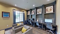 This single-floor community space is thoughtfully designed to serve as both a business center and a social lounge, seamlessly blending functionality with inviting aesthetics. The interior is finished with warm yellow-toned walls, recessed lighting, and wood-look flooring throughout, creating a cohesive and welcoming atmosphere.

The dedicated workstation area features a built-in dark-toned shelving and desk unit that spans an entire wall, accommodating multiple computer workstations side by side. Ergonomic adjustable chairs are positioned at each station, and the upper shelves are styled with bold, abstract circular artwork, adding a contemporary artistic touch. A large window with striped drapery panels allows natural light to brighten the workspace.

Adjacent to the business area, the lounge space offers a comfortable seating arrangement with upholstered gray armchairs accented by yellow throw pillows, gathered around a rustic wood coffee table atop a patterned area rug. Elegant table lamps flank a decorative console, and additional artwork enhances the walls. A set of sliding barn-style glass doors with a dark hardware track connects the lounge to adjacent areas, providing both visual openness and flexible separation. The overall design strikes a balance between professional utility and relaxed comfort.