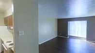 This single-floor space offers a clean and straightforward layout designed for functional living. The main living area is defined by rich, dark hardwood flooring that runs throughout, providing a strong visual contrast against the light-colored walls. The walls feature a two-tone design, with soft cream tones on the primary surfaces and a deeper, darker accent wall near the window, adding depth and visual interest to the room. A textured popcorn ceiling adds a classic touch overhead. Natural light filters into the space through a large window dressed with vertical blinds, brightening the interior and creating an open, airy feel. White baseboards cleanly frame the perimeter of the room, offering a crisp finishing detail. Electrical outlets are positioned along the walls for practical use throughout the space. The room's open feel and simple, neutral palette make it a versatile canvas well-suited for a variety of furnishing styles and interior arrangements.