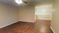 This single-story space offers a clean, functional layout with warm hardwood-style flooring that runs continuously throughout the main living and dining areas, creating a cohesive and inviting atmosphere. The rich, dark tones of the flooring contrast attractively against the neutral beige walls and crisp white baseboards, lending the space a polished yet understated aesthetic.

The main living area is outfitted with a ceiling fan featuring a multi-bulb light fixture, providing both ventilation and ambient lighting. The walls are smooth and neutrally painted, offering a versatile backdrop suitable for a variety of interior styles and furnishings.

An open passage connects the living area to an adjacent dining space, which is distinguished by a pendant light fixture that hangs centrally, providing focused task lighting over a dining arrangement. The transition between the two areas is defined by a subtle change in wall height, adding a modest architectural detail to the otherwise straightforward layout.

A door visible from the main living area suggests access to additional rooms or storage. White baseboards throughout provide clean, classic finishing detail. Overall, the space presents a tidy, move-in-ready environment with durable finishes well-suited for everyday living.