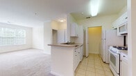 Located at 9901 Gable Ridge Terrace in Rockville, Maryland, this single-floor residence offers a well-organized layout designed for comfortable everyday living. The kitchen is outfitted with white cabinetry, light-toned countertops, and a full suite of white appliances including a refrigerator, gas range, and built-in microwave. A peninsula with an integrated sink and breakfast bar seamlessly connects the kitchen to the adjacent dining and living areas, both finished with neutral wall-to-wall carpeting and crown molding details that lend a polished feel throughout. The living room is generously proportioned, featuring large windows with blinds that invite abundant natural light and offer views of the surrounding greenery, along with a glass door providing access to an outdoor area. A bedroom with soft carpeting and clean, neutral walls offers a serene retreat, complemented by good closet space with paneled doors. The full bathroom is finished with light tile flooring, a white vanity with under-cabinet storage, a mirror, and a full shower-tub combination. Light tile flooring continues through the kitchen and bathroom areas, providing a practical contrast to the carpeted living spaces. The overall palette of soft whites and warm neutrals creates a bright, airy ambiance throughout the home.
