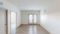 This single-floor unit presents a clean, modern aesthetic defined by a neutral white color palette, light wood-tone finishes, and an efficient layout. The kitchen is a standout feature, equipped with a full suite of stainless steel appliances — including a refrigerator, electric range, microwave, and dishwasher — complemented by contemporary flat-front cabinetry in a warm wood finish, light quartz-style countertops, and a white subway tile backsplash. Light-toned luxury vinyl plank flooring extends throughout the kitchen and main living areas, contributing to a cohesive and easy-to-maintain interior.

The living area is bright and open, anchored by a set of French doors fitted with horizontal blinds that allow for natural light while offering privacy. The bedroom areas feature soft carpeting and are well-lit by windows that provide outdoor views. The bathroom is finished with matching wood-toned cabinetry, a single-basin vanity with a light countertop, wood-look vinyl flooring, and a full tub/shower combination with brushed nickel fixtures. Recessed lighting throughout reinforces the clean, contemporary character of the space.
