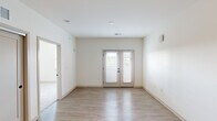 This single-floor unit presents a clean, modern aesthetic defined by a neutral white color palette, light wood-tone finishes, and an efficient layout. The kitchen is a standout feature, equipped with a full suite of stainless steel appliances — including a refrigerator, electric range, microwave, and dishwasher — complemented by contemporary flat-front cabinetry in a warm wood finish, light quartz-style countertops, and a white subway tile backsplash. Light-toned luxury vinyl plank flooring extends throughout the kitchen and main living areas, contributing to a cohesive and easy-to-maintain interior.

The living area is bright and open, anchored by a set of French doors fitted with horizontal blinds that allow for natural light while offering privacy. The bedroom areas feature soft carpeting and are well-lit by windows that provide outdoor views. The bathroom is finished with matching wood-toned cabinetry, a single-basin vanity with a light countertop, wood-look vinyl flooring, and a full tub/shower combination with brushed nickel fixtures. Recessed lighting throughout reinforces the clean, contemporary character of the space.